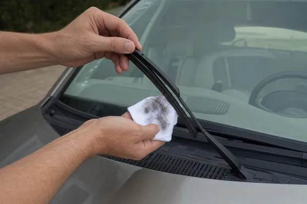One hand lifting a wiper arm off the windscreen, the other wiping the rubber edge with a damp cloth that shows a dark smear